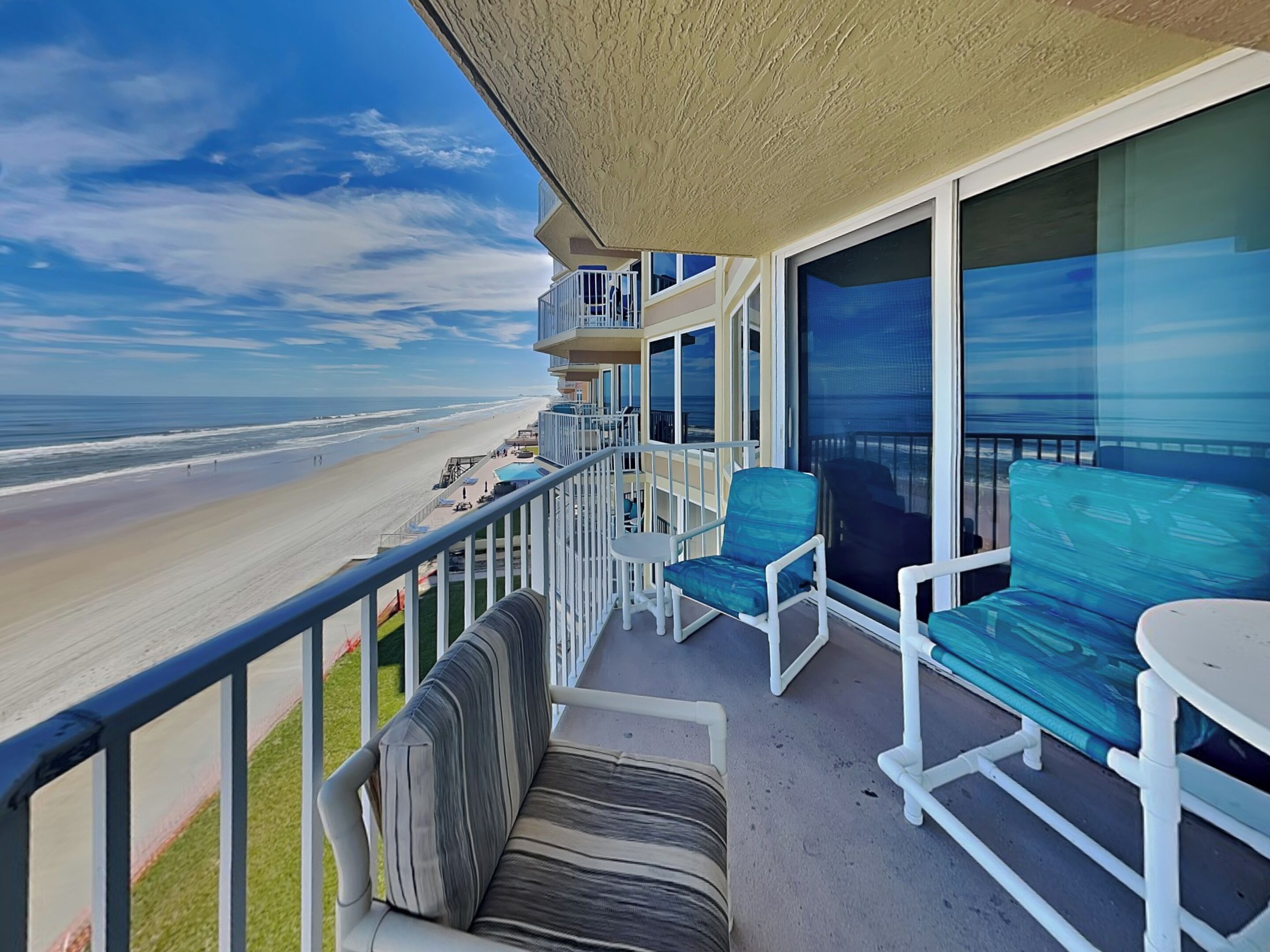 A balcony with a view of the beach, a pool, and a seating area with chairs and a sofa.