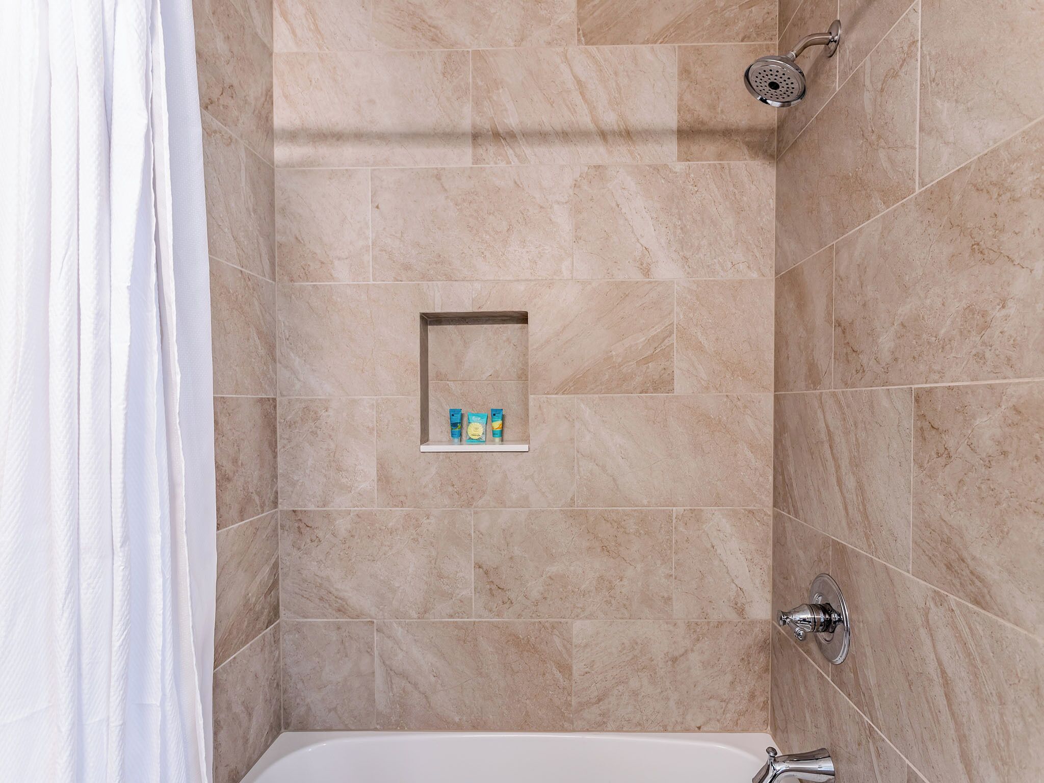 Un baño con azulejos color crema, una ducha, un estante empotrado con artículos de aseo y una bañera blanca.