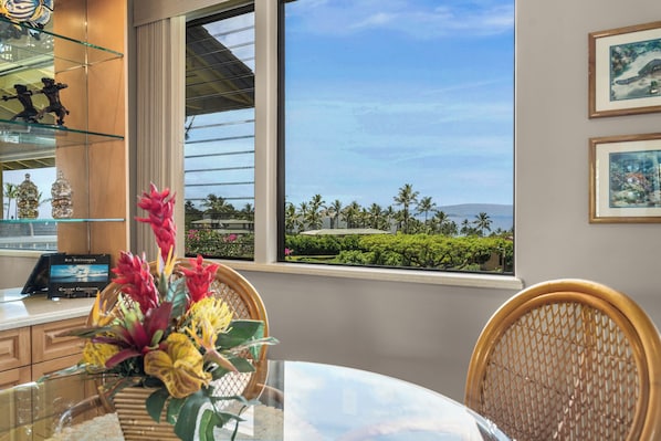 A hotel room with a view of palm trees and mountains, a glass table with a flower arrangement, and a wooden chair. - Wailea Ekahi by Maui Rental Group (Kihei)