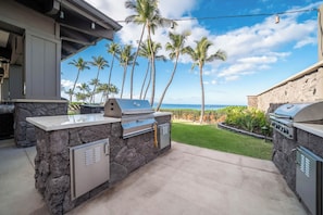An outdoor kitchen with a stone countertop, a built-in grill, and a view of the ocean and palm trees. - Wailea Ekahi by Maui Rental Group (Kihei)
