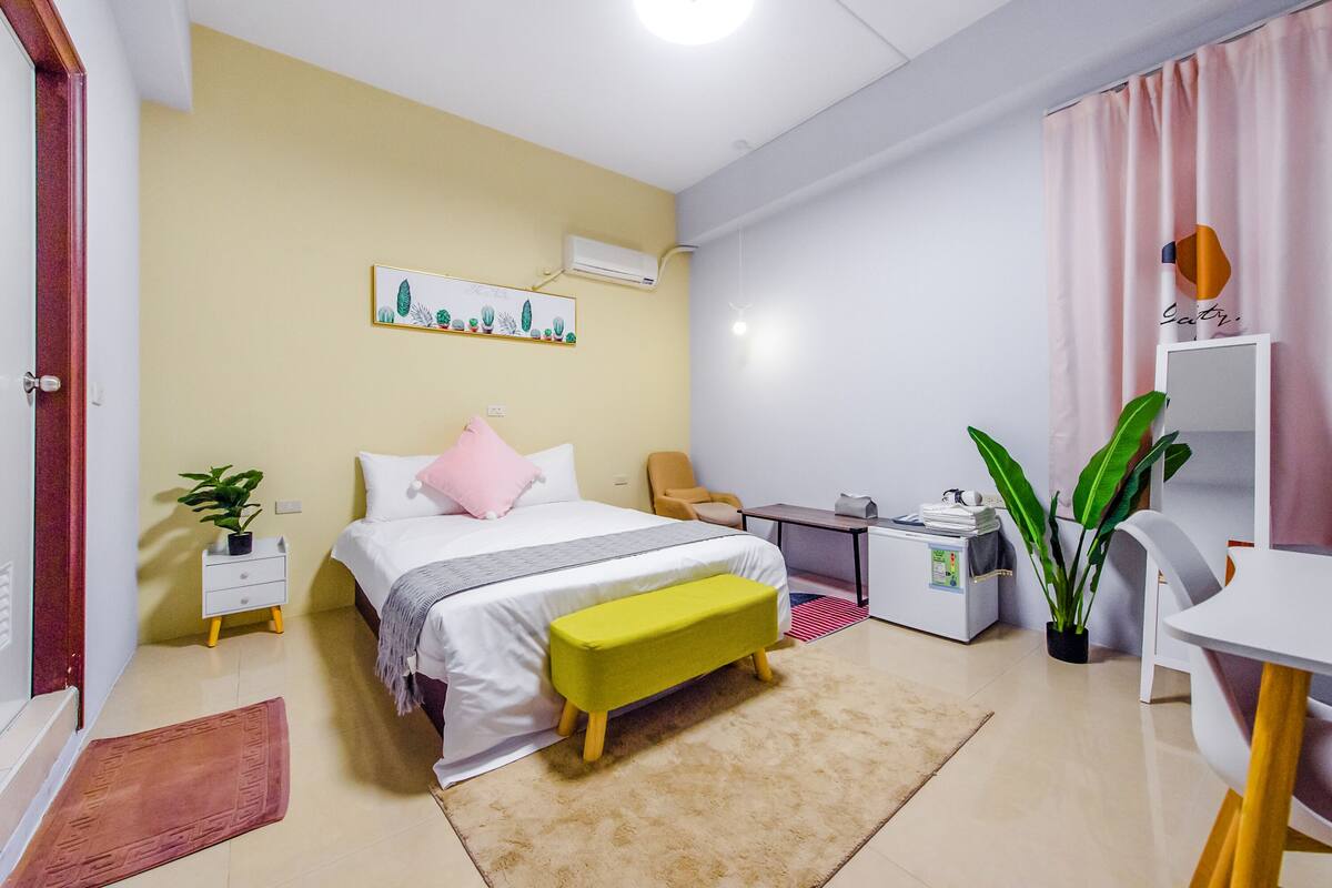 A bedroom with a bed, bedside table, chair, desk, and a small kitchenette.