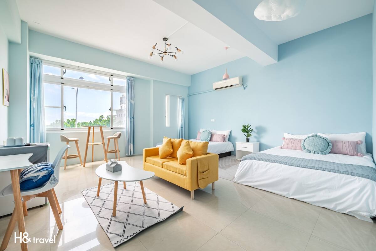 A modern hotel room with a yellow sofa, a white bed, and a dining area with a table and chairs.