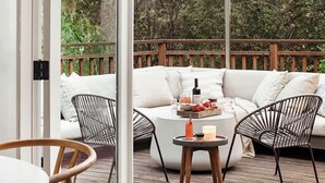 An outdoor deck with a wooden floor, a white sofa with cushions, two wire chairs, a round table with a tray of food and drinks, and a hanging light fixture. - THE Zen Treehouse Retreat- Great Views and Peaceful Decks!
 (Los Angeles)