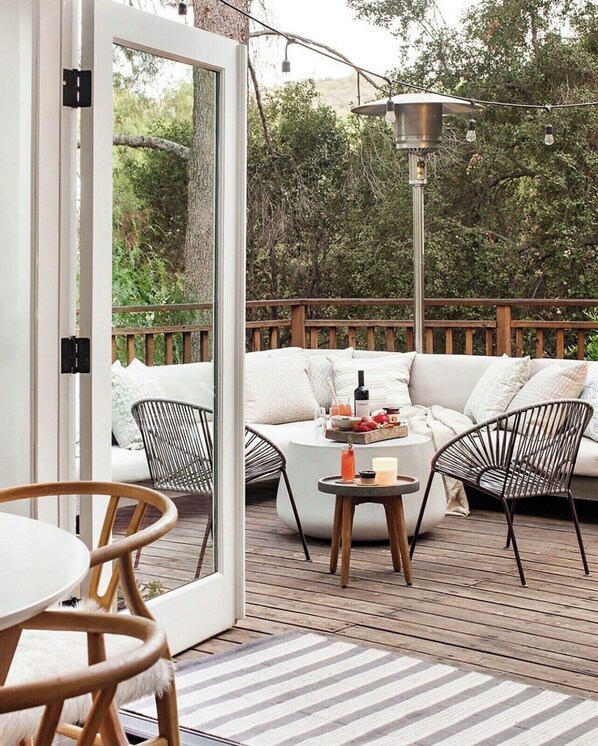 An outdoor deck with a wooden floor, a white sofa with cushions, two wire chairs, a round table with a tray of food and drinks, and a hanging light fixture. - THE Zen Treehouse Retreat- Great Views and Peaceful Decks!
(Los Angeles)