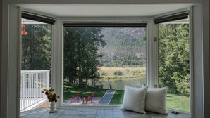 A room with a large window offering a view of a lake, trees, and a grassy area. - All-Season Family Retreat • Hot Tub • Waterfront (Moyie)