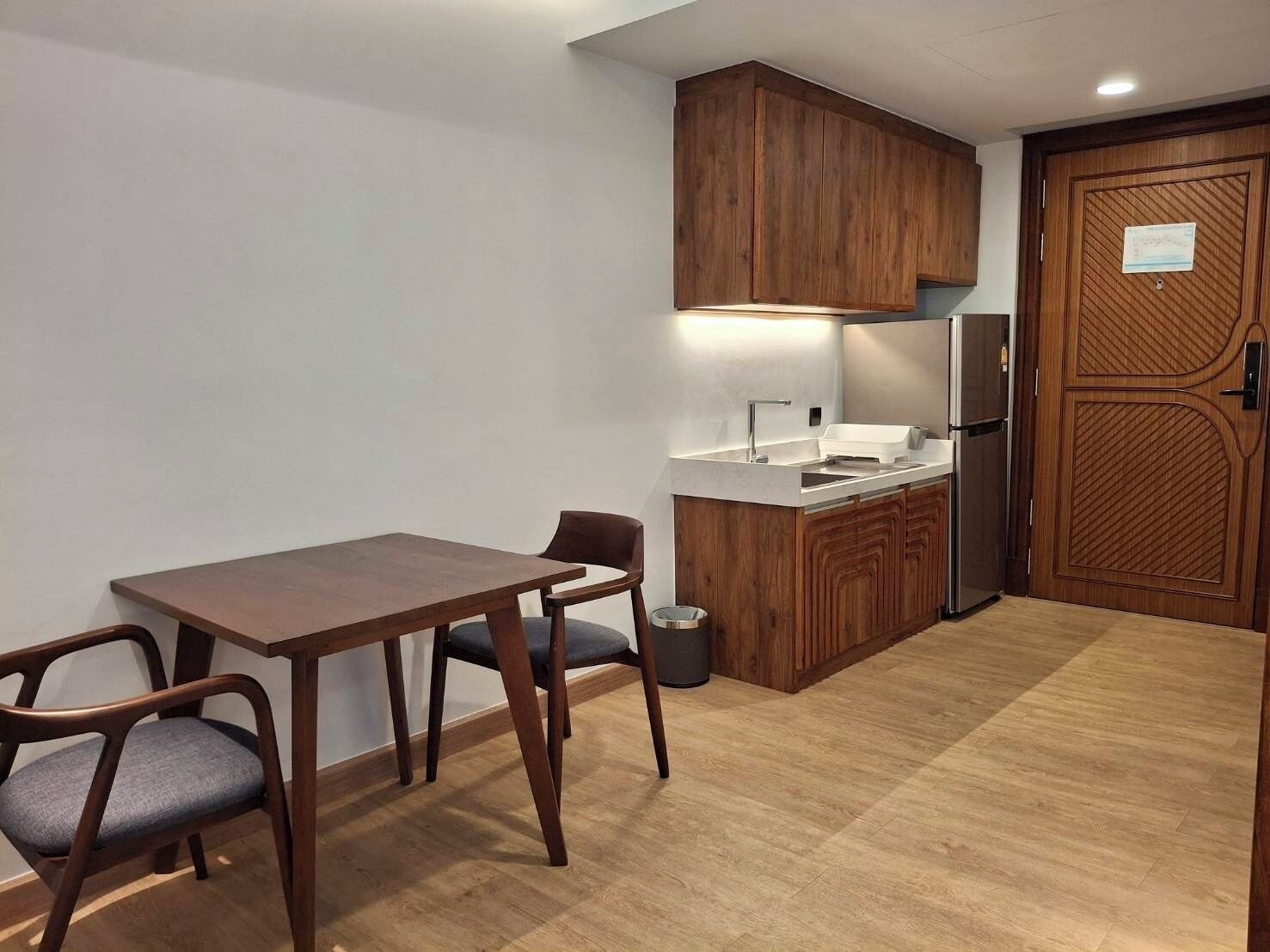 Una cocina compacta con armarios de madera, un refrigerador de acero inoxidable, una encimera blanca y una mesa de comedor con dos sillas.