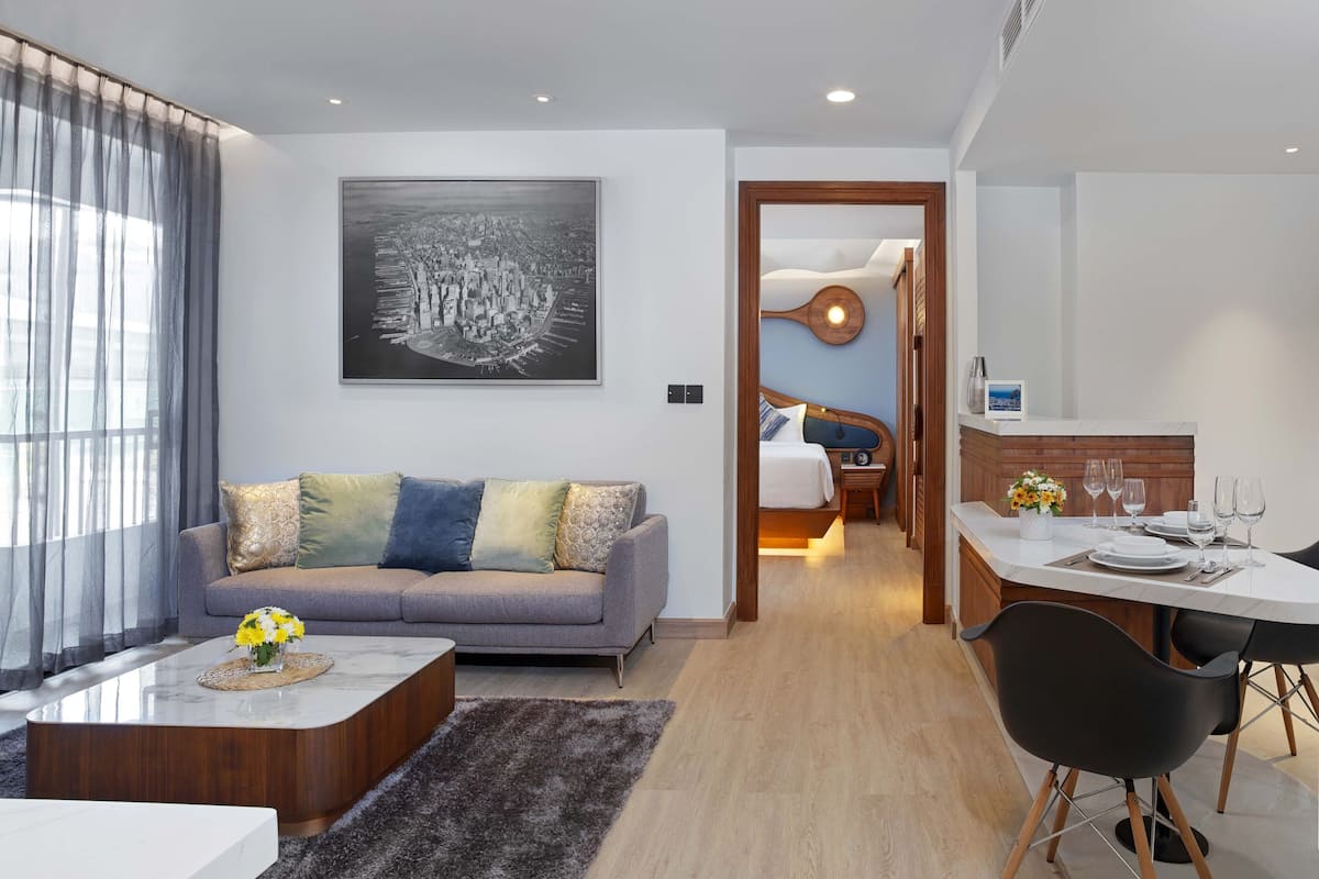 A modern hotel room with a grey sofa, a dining table set for two, and a large framed cityscape artwork on the wall.