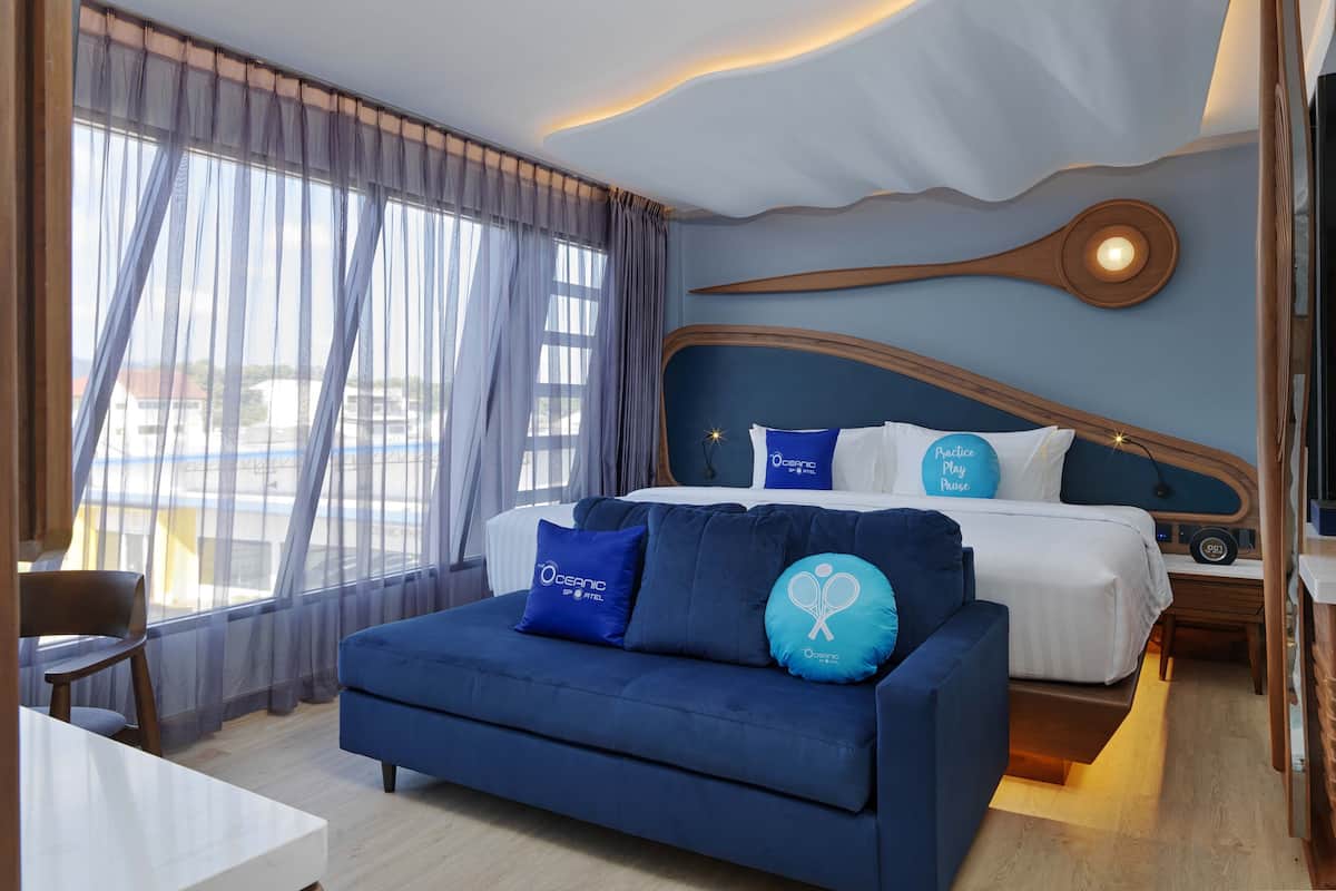 A hotel room with a bed, a blue sofa, a wooden headboard, and a view of the outside.