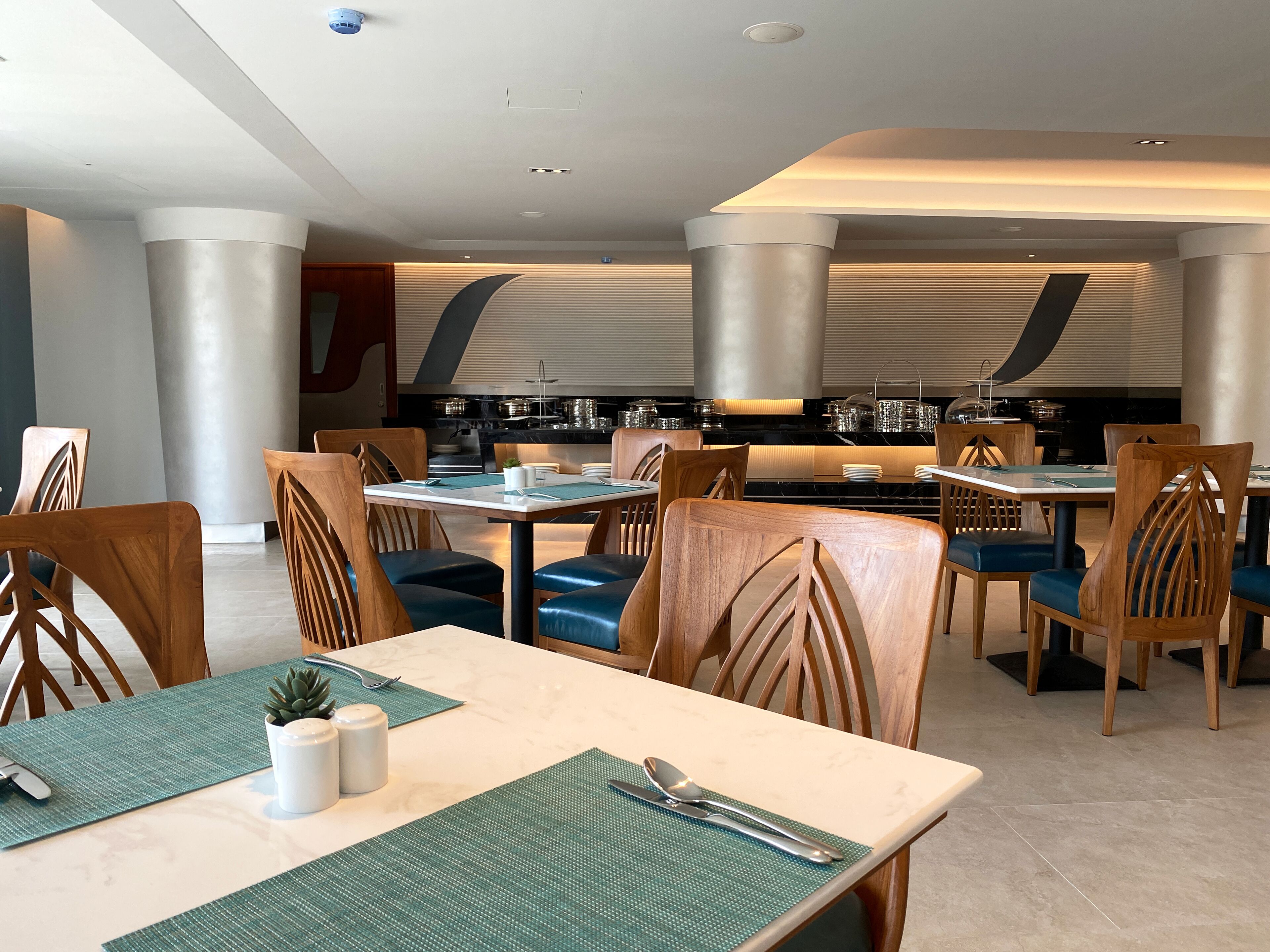 A modern restaurant with wooden chairs and tables, a bar area, and a kitchen in the background.