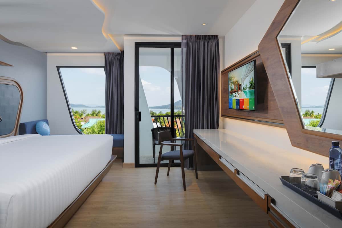 A modern hotel room with a bed, a desk, a chair, a TV, and a balcony with a view of the sea.