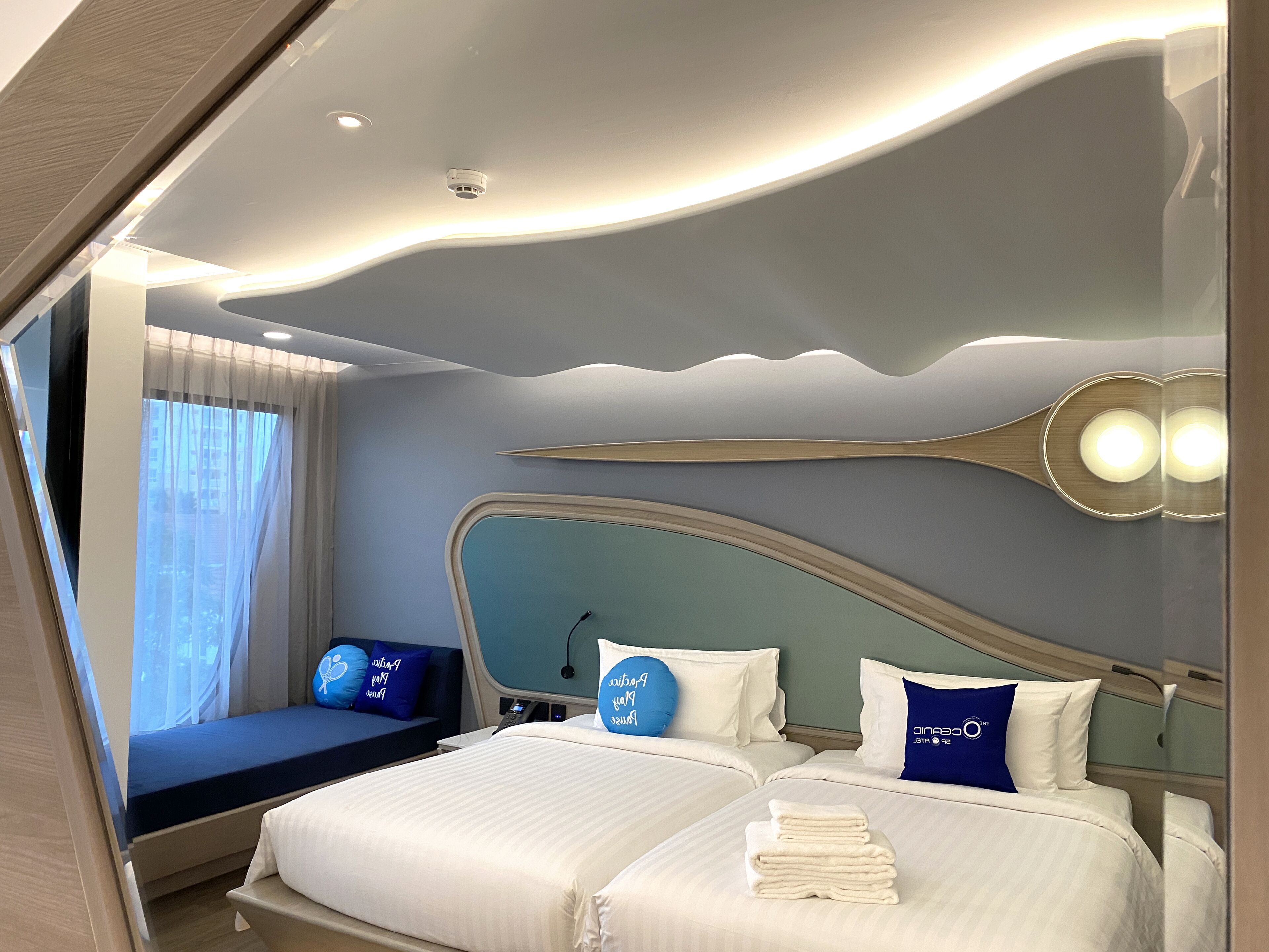 Una camera d'albergo moderna con soffitto curvo, due letti con lenzuola bianche e azzurre, un comodino con una lampada e vista all'esterno attraverso una finestra con tende leggere.