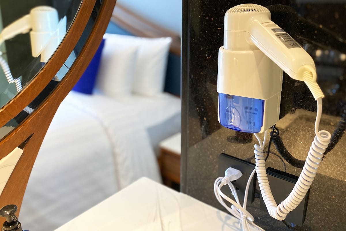 A hairdryer on a bathroom counter with a mirror reflecting a bed and a nightstand.