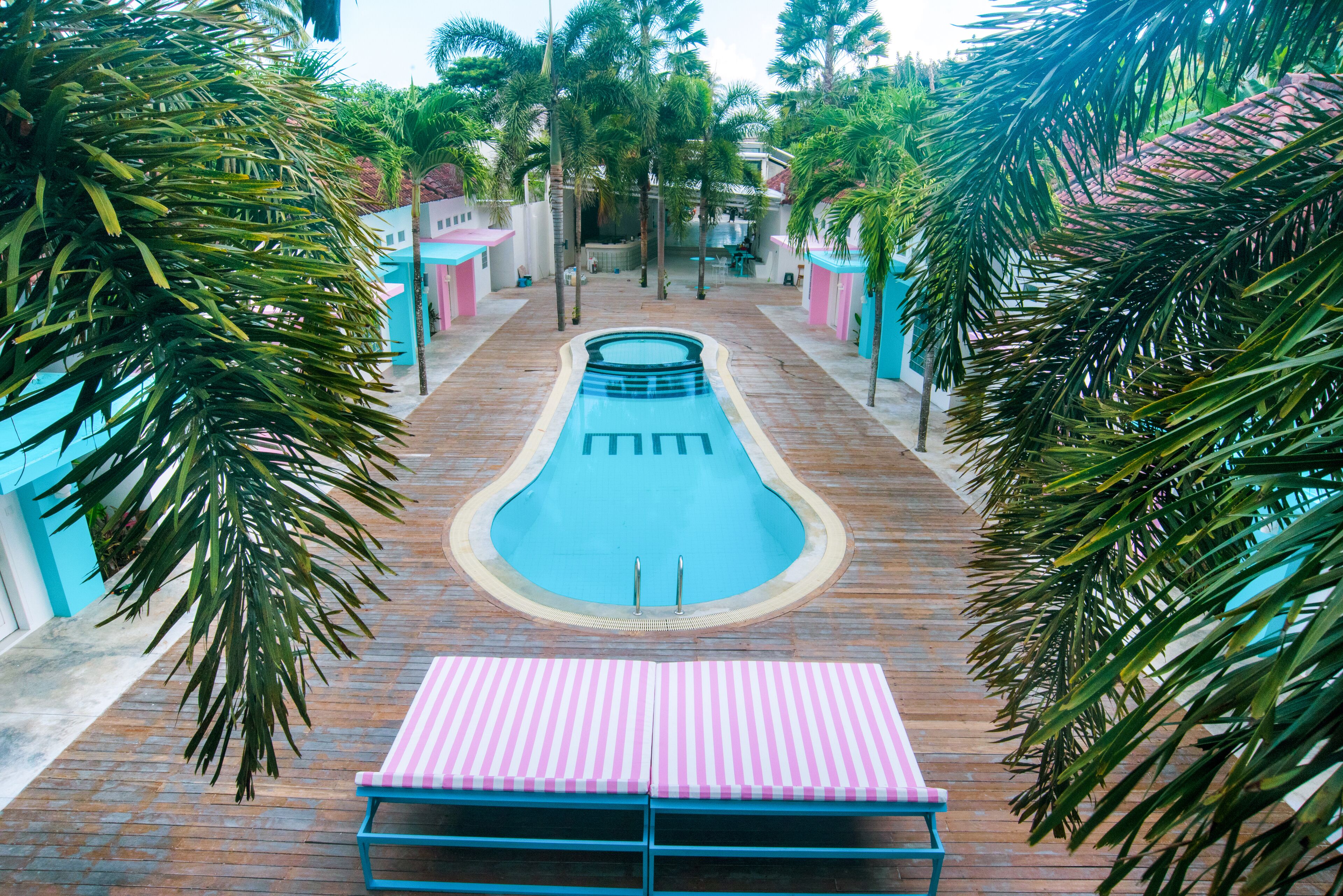 Zwembad met een loungehoek in roze en blauw tinten op de voorgrond, omgeven door palmbomen en gebouwen met pastelkleurige gevels.