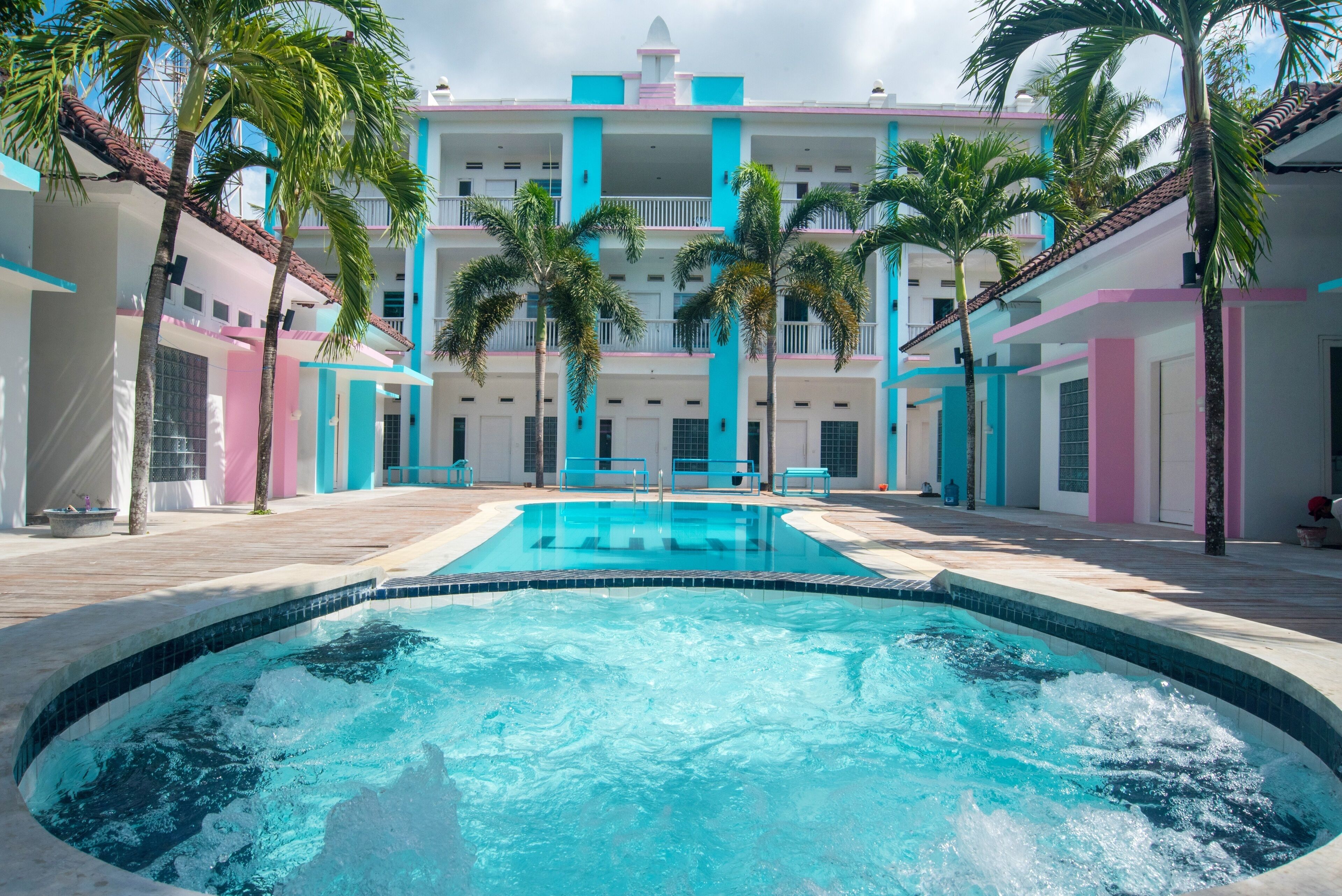 A swimming pool surrounded by a multi-story building with balconies, palm trees, and a tiled deck.