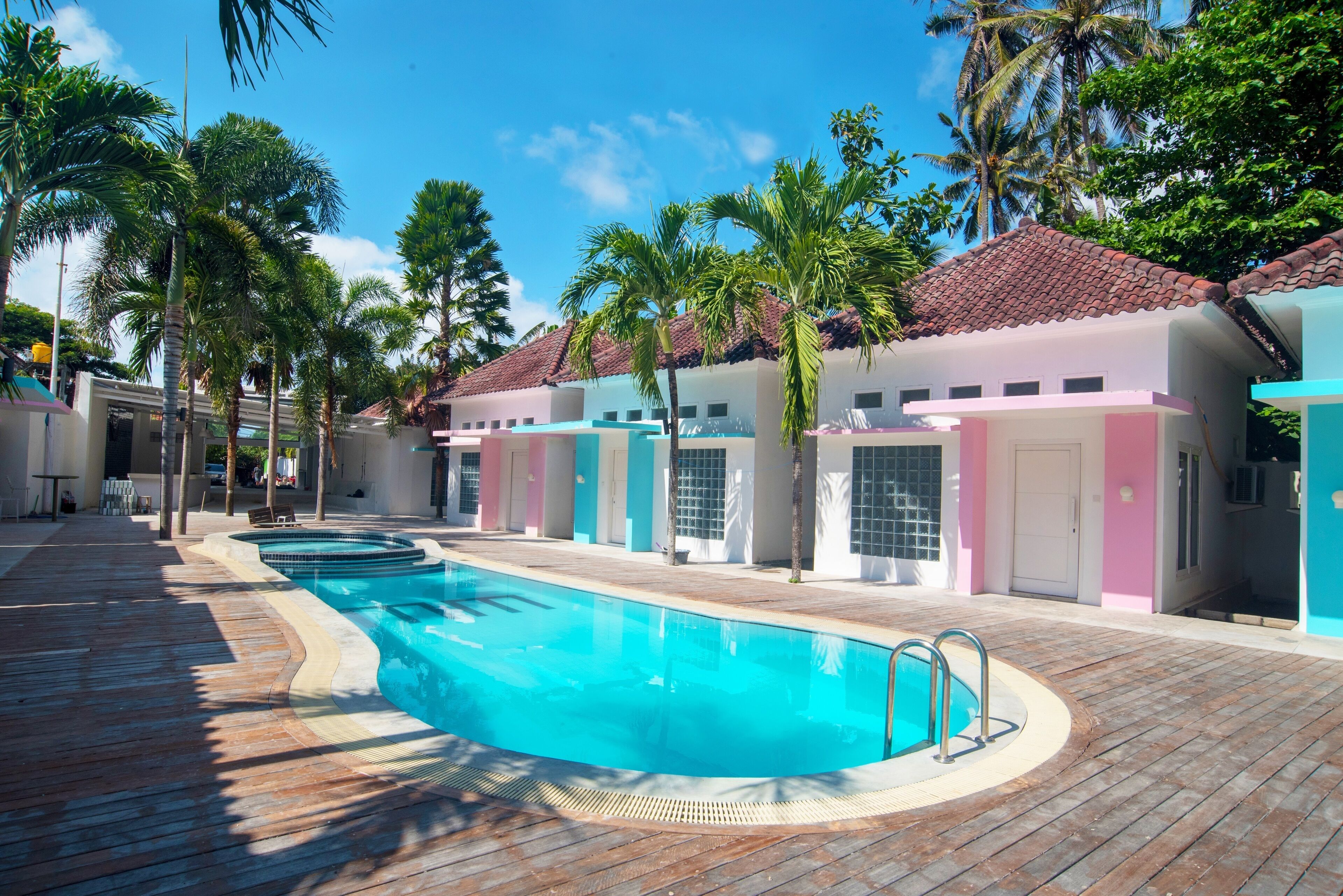 A swimming pool with a wooden deck, surrounded by palm trees and buildings with pastel-colored facades.