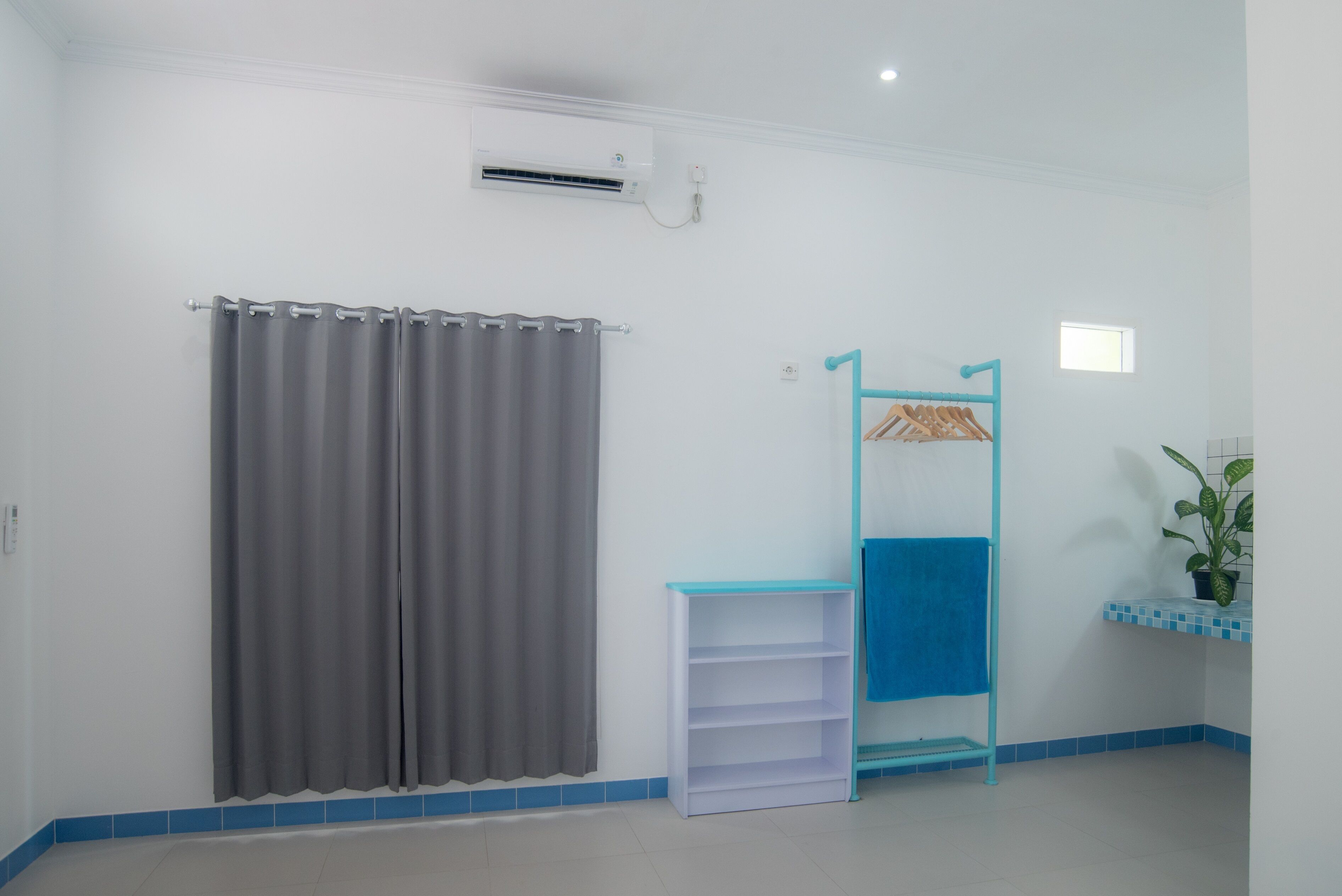 Een badkamer met een grijs gordijn, een witte airconditioning, een blauwe handdoekhouder, een wit plankje en een klein raam.