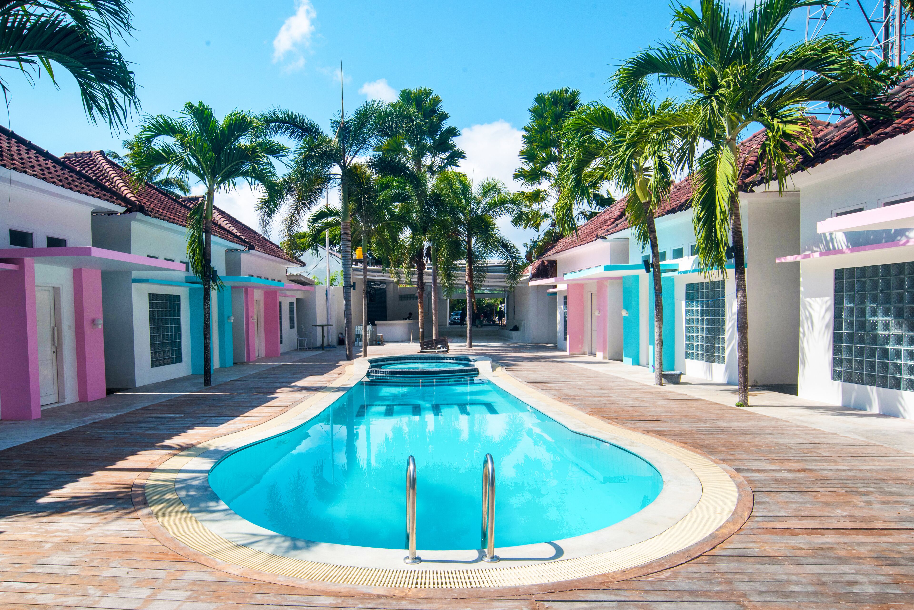 A swimming pool with a wooden deck, surrounded by palm trees and buildings with pastel-colored facades.