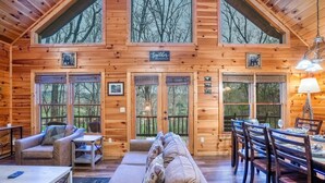 A log cabin interior with a dining area, a sofa, and a large window overlooking trees. - ★Cozy Cabin with Hot Tub, Game Room, and Fire pit!★ Wears Valley! (Sevierville)