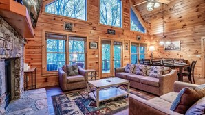 A cozy cabin interior with a stone fireplace, wooden walls, large windows, and a dining area. - ★Cozy Cabin with Hot Tub, Game Room, and Fire pit!★ Wears Valley! (Sevierville)