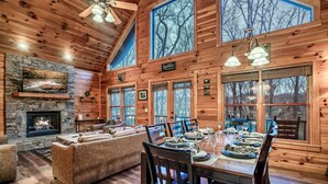 A log cabin interior with a stone fireplace, a dining area set for six, and a large window overlooking trees. - ★Cozy Cabin with Hot Tub, Game Room, and Fire pit!★ Wears Valley! (Sevierville)