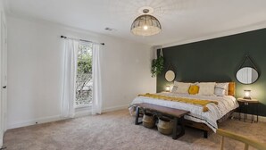 Ein Schlafzimmer mit Bett, Nachttischen, einer Bank, einer Hängelampe und Blick durch ein mit Vorhängen drapiertes Fenster.