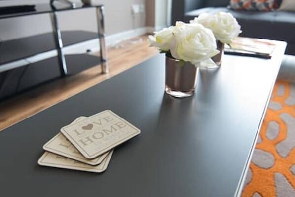 A modern living room with a dark wooden coffee table, a vase with white flowers, and a stack of coasters with the text "I LOVE HOME".