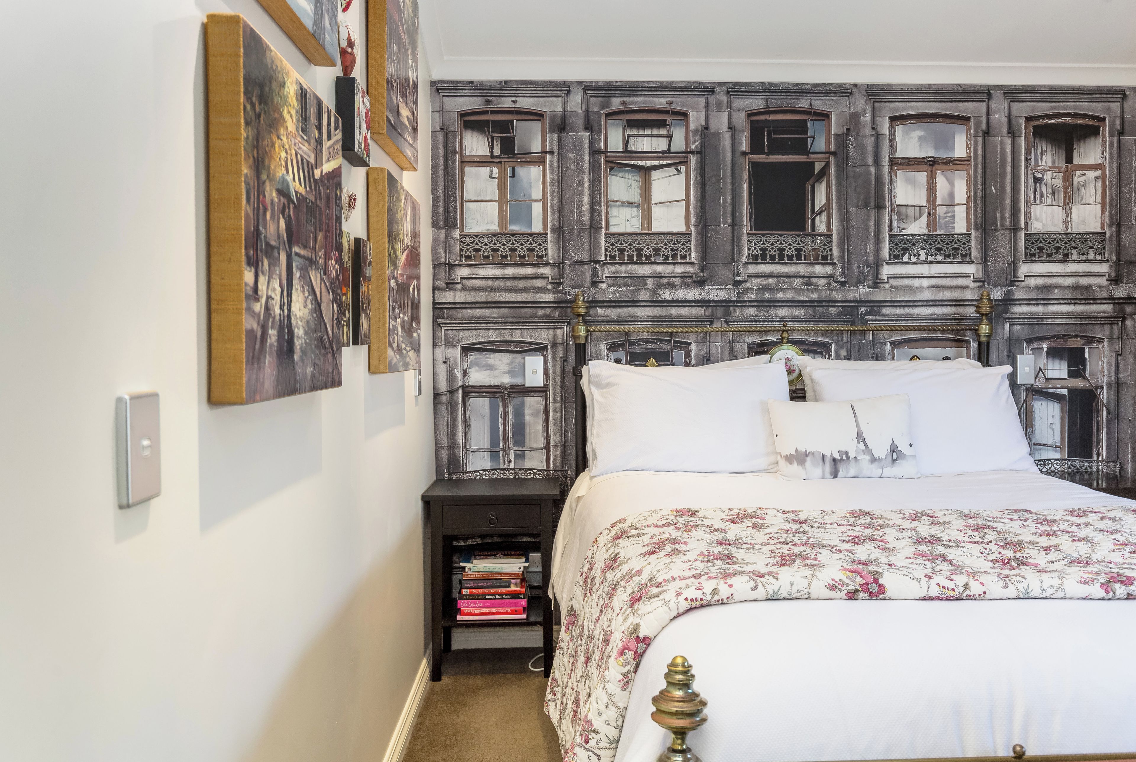 Quarto com uma cama, mesa de cabeceira com livros e parede decorada com quadros.