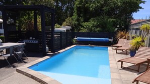 Een rechthoekig zwembad met een blauw binnenoppervlak, omringd door een betegeld zonnedek. Er staan houten ligbedden en er is een barbecueplaats met een zwart dakzeil.