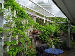 Een overdekt terras met houten omheining, witte bloemen die hangen, en een tafel met stoelen.
