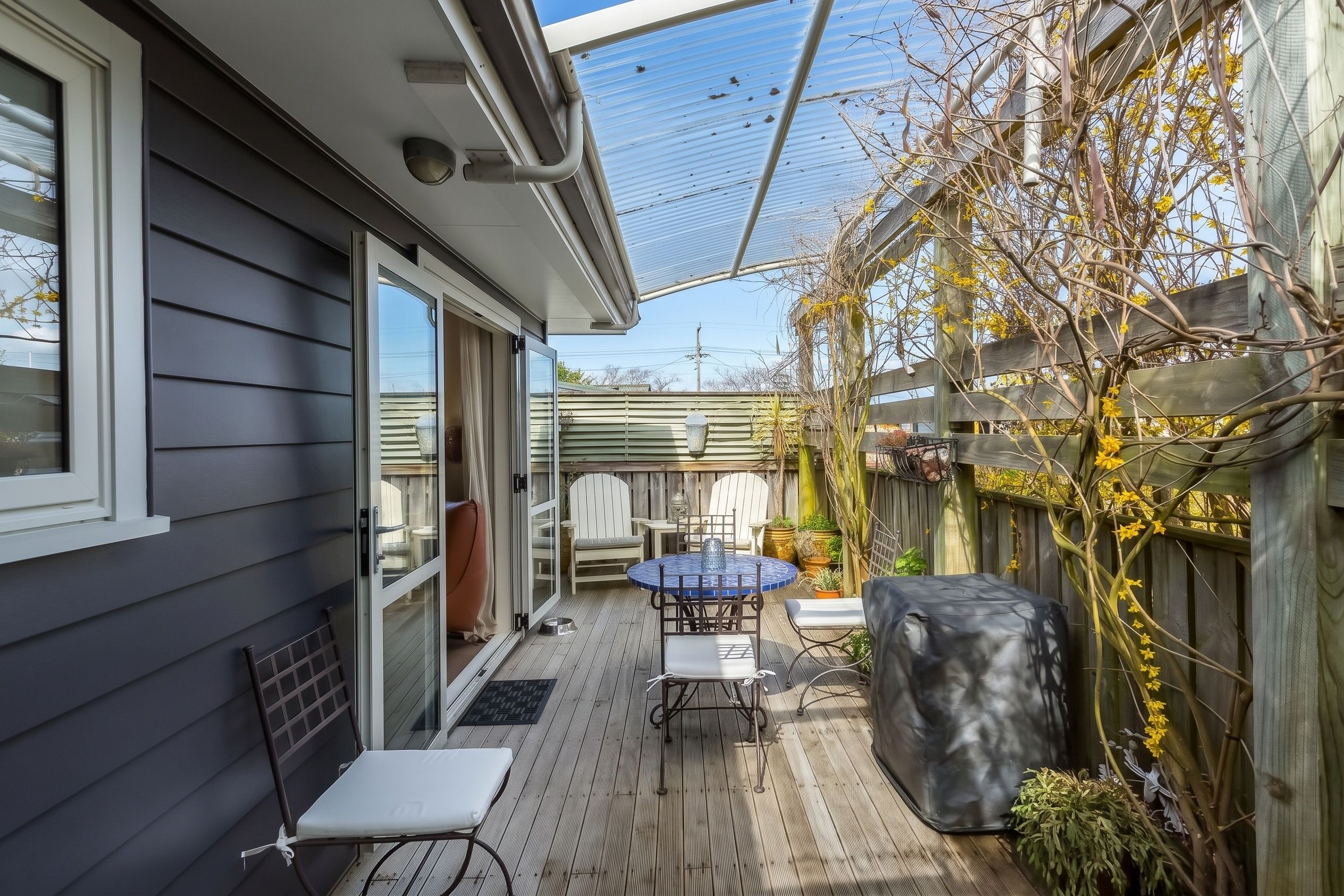 Eine überdachte Terrasse mit Tisch und Stühlen, einem Grill und einem Holzzaun mit Kletterpflanzen.