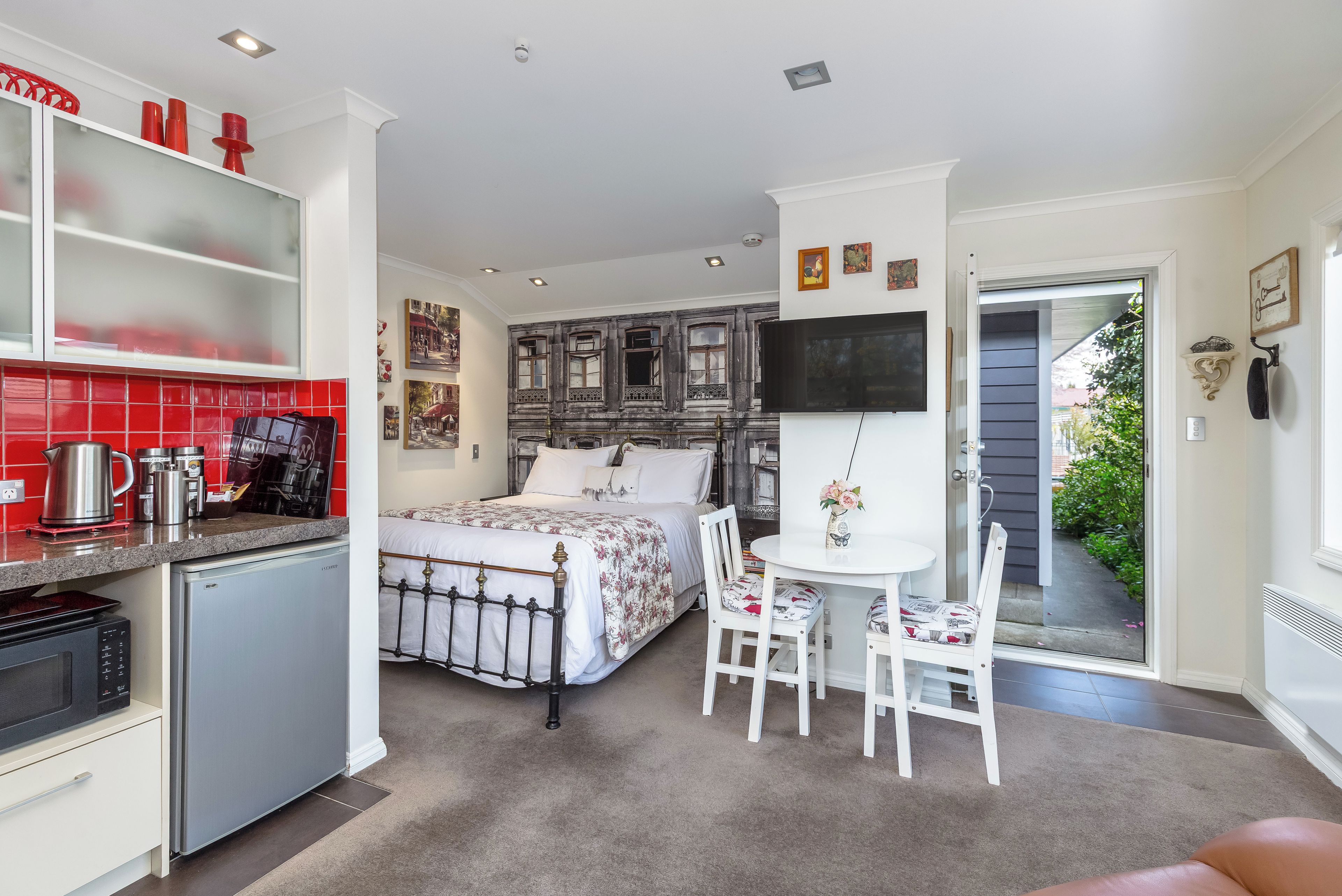 A compact hotel room with a kitchenette, a bed with a floral bedspread, a dining area with a table and chairs, and a TV mounted on the wall.