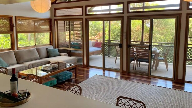 Amplia sala de estar con sofá, mesa de centro y mesa de comedor. Grandes puertas correderas de vidrio dan acceso a un patio exterior con terraza de madera y mobiliario.