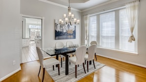 A dining area with a glass table, white chairs, a chandelier, and a cityscape painting. - Big 5 Bed/ 3.5 Bath Modern Home ⌂ Disney+ included, Free Parking, Fast Wi-Fi (Plano)