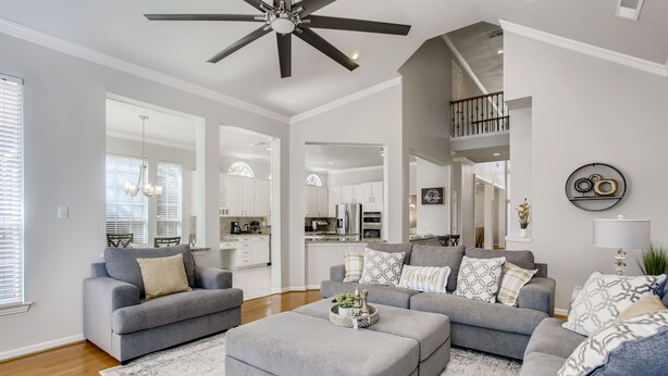 A spacious living room with grey sofas and a large ottoman, a ceiling fan, and a view into the kitchen. - Big 5 Bed/ 3.5 Bath Modern Home ⌂ Disney+ included, Free Parking, Fast Wi-Fi (Plano)