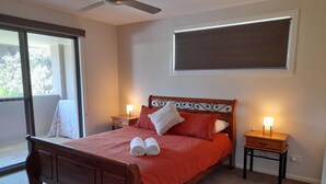 A neatly made bed with a wooden headboard, two bedside tables with lamps, a large window with a view of greenery, and a ceiling fan. - 'SEASIDE RETREAT' beach & nature at your doorstep (Wooli)