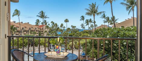 A balcony with a round table and two chairs, overlooking a pool area with palm trees and a clear blue sky.