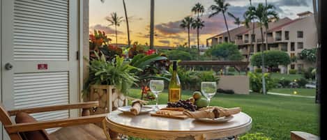 A wooden table with a bottle, glasses, and snacks on a balcony overlooking a garden with palm trees and a building in the background.