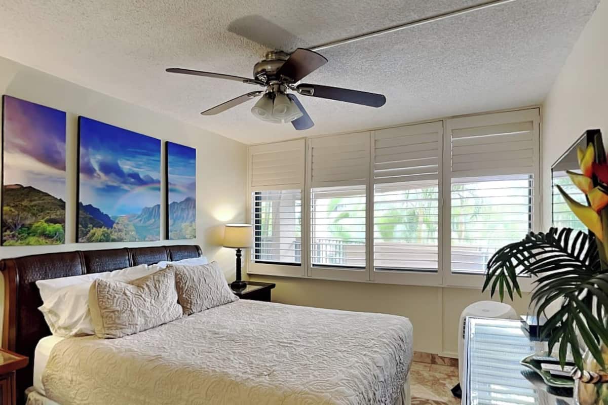 Ein Schlafzimmer mit einem Bett, einem Deckenventilator, einem Fenster mit Jalousien und einem Nachttisch.