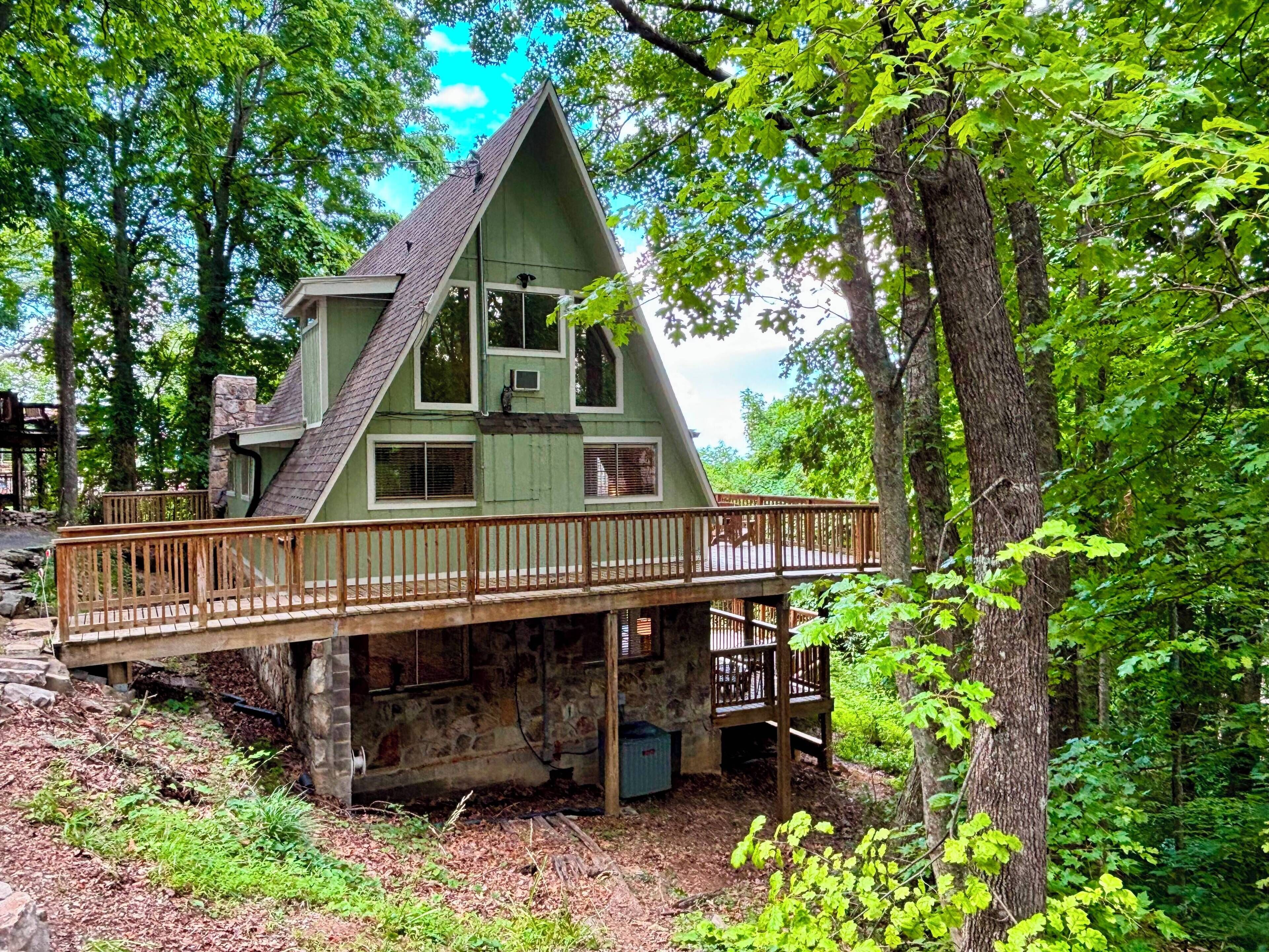 A house with a wooden deck and a stone foundation, surrounded by trees.