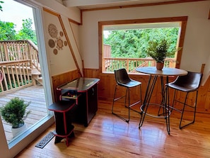 Una habitación con piso de madera, una mesa de vidrio con dos sillas, una mesita con una sola silla, una terraza de madera afuera y una ventana con vista a vegetación.