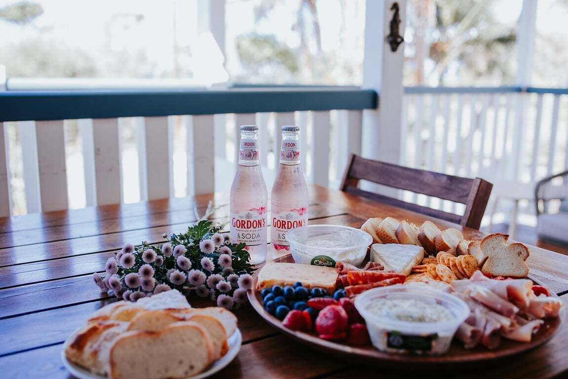 Une table en bois sur laquelle sont disposés divers aliments, notamment du pain, du fromage et des fruits, ainsi que deux bouteilles de soda de la marque Gordon's.