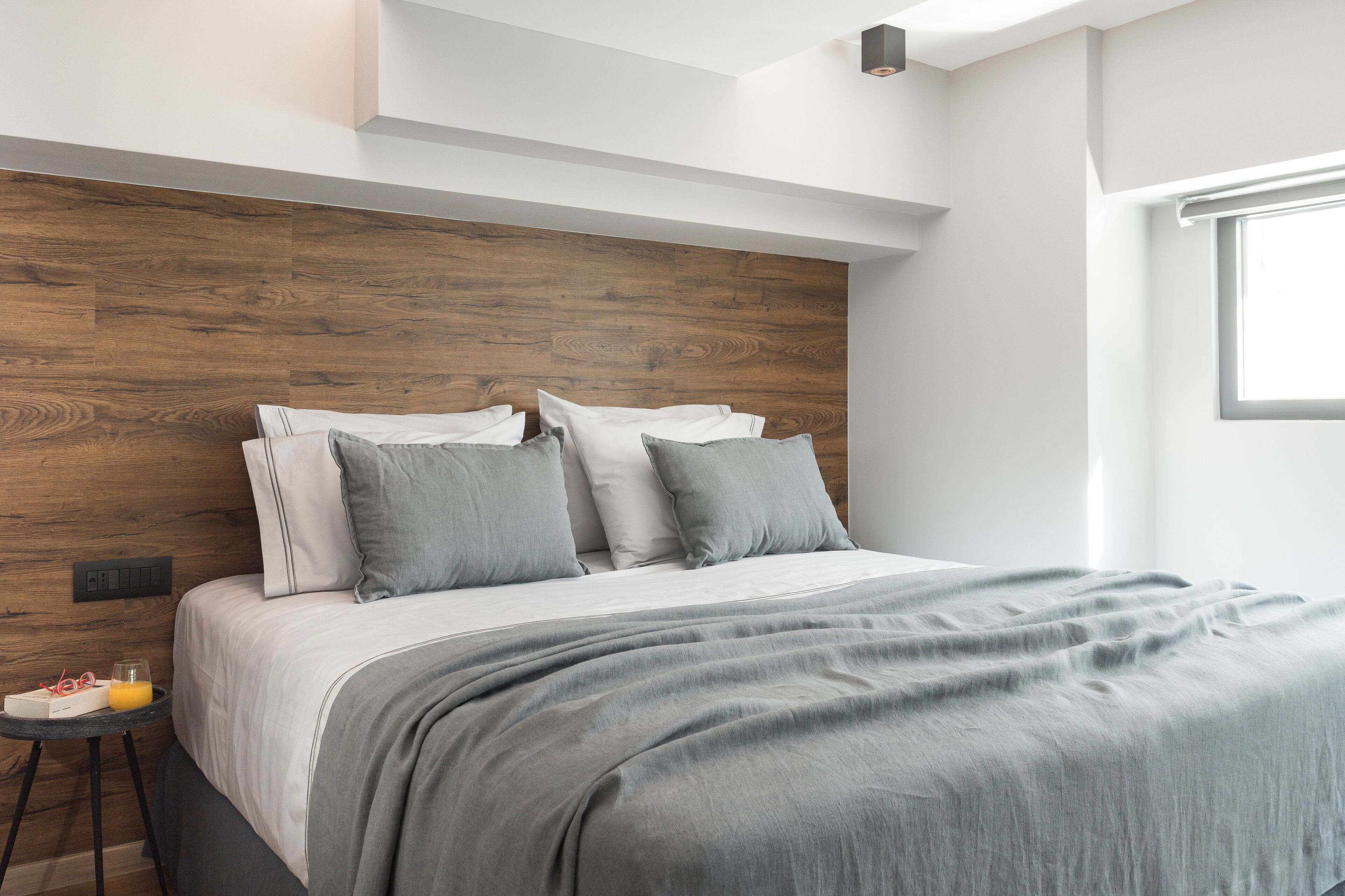 A modern bedroom with a wooden headboard, a neatly made bed with grey bedding, and a small bedside table with a glass of orange juice.
