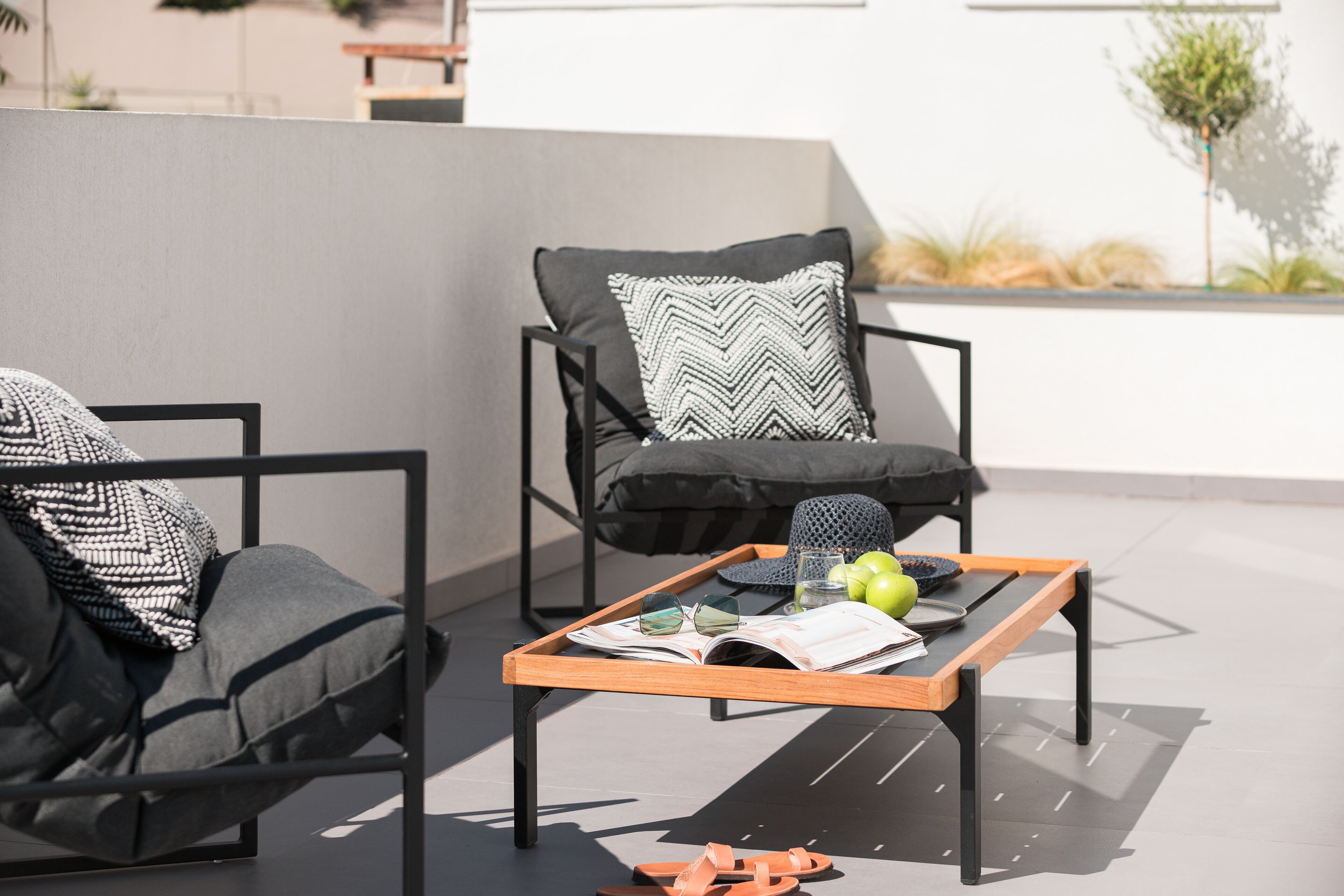 A modern outdoor seating area with two chairs, a wooden table, and a tray with books, glasses, and apples.