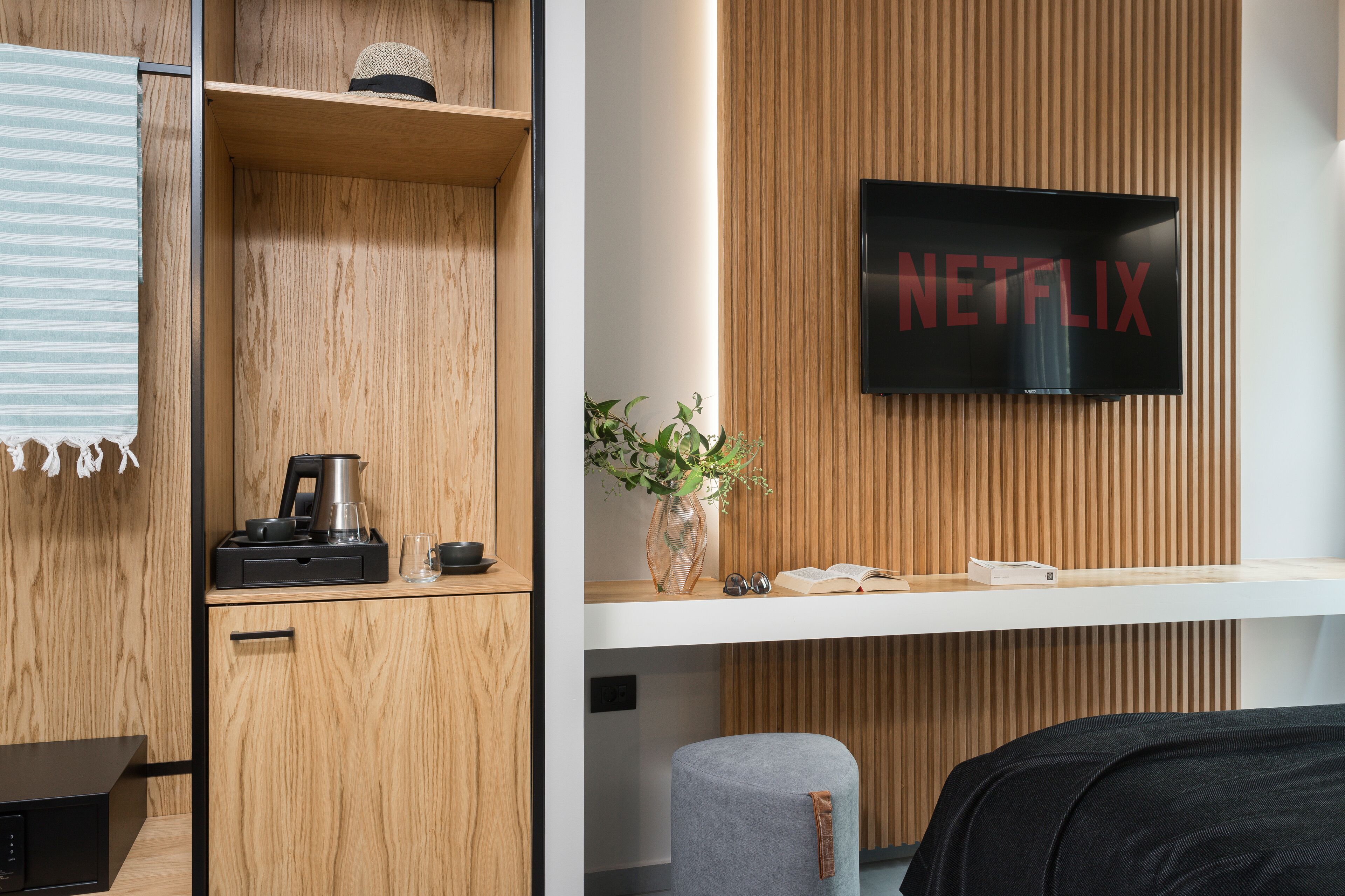 A modern hotel room with wooden paneling, a flat-screen TV, a coffee maker, a vase with a plant, and a bedside table with a book and glasses.