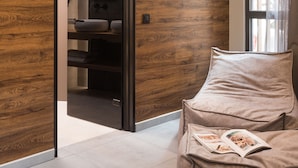 Une salle de bain moderne avec un mur en bois, une cabine de douche en verre et un coin salon beige avec des coussins et un magazine.