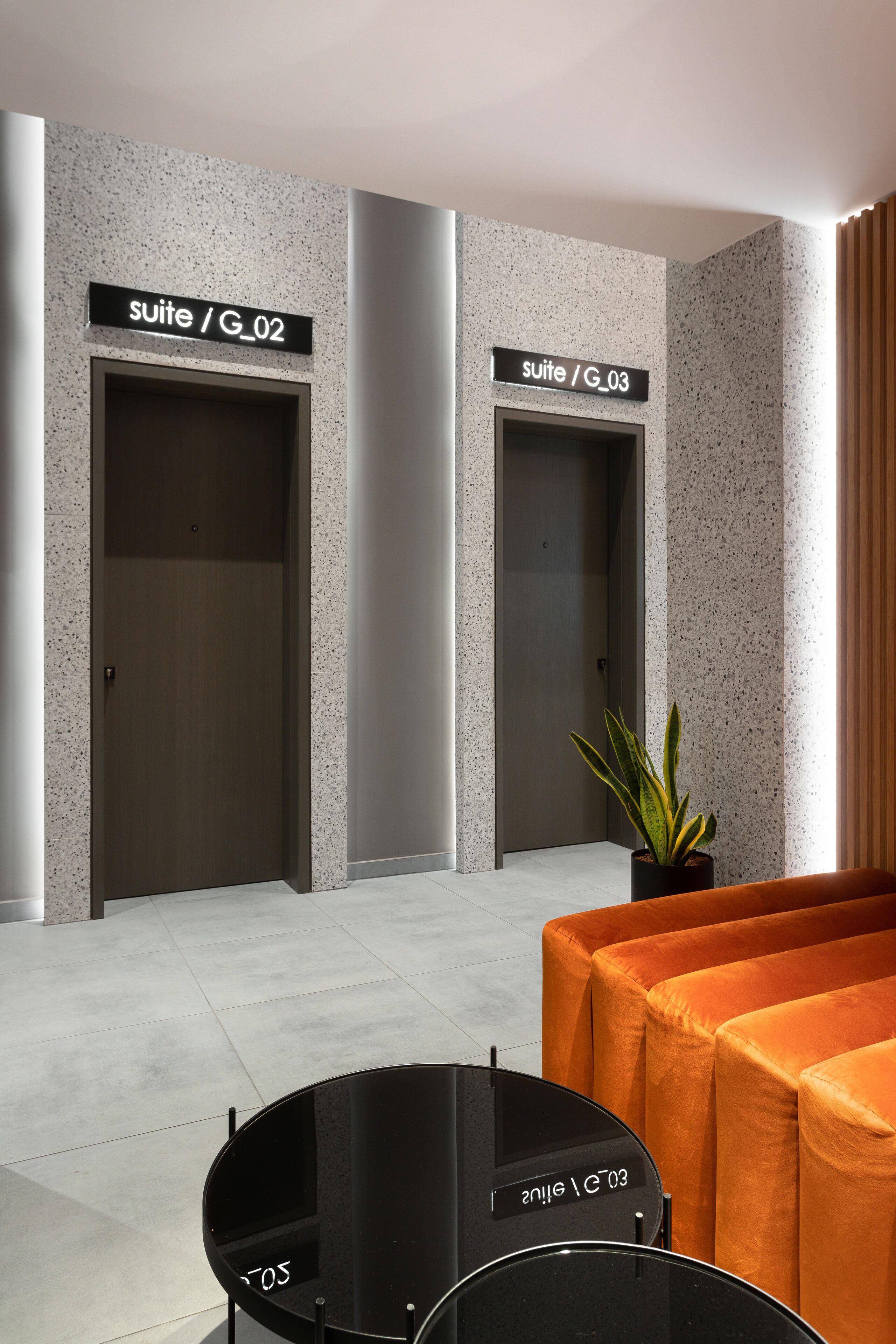 A modern lobby with two elevators marked "suite / G_02" and "suite / G_03", a potted plant, and an orange sofa.