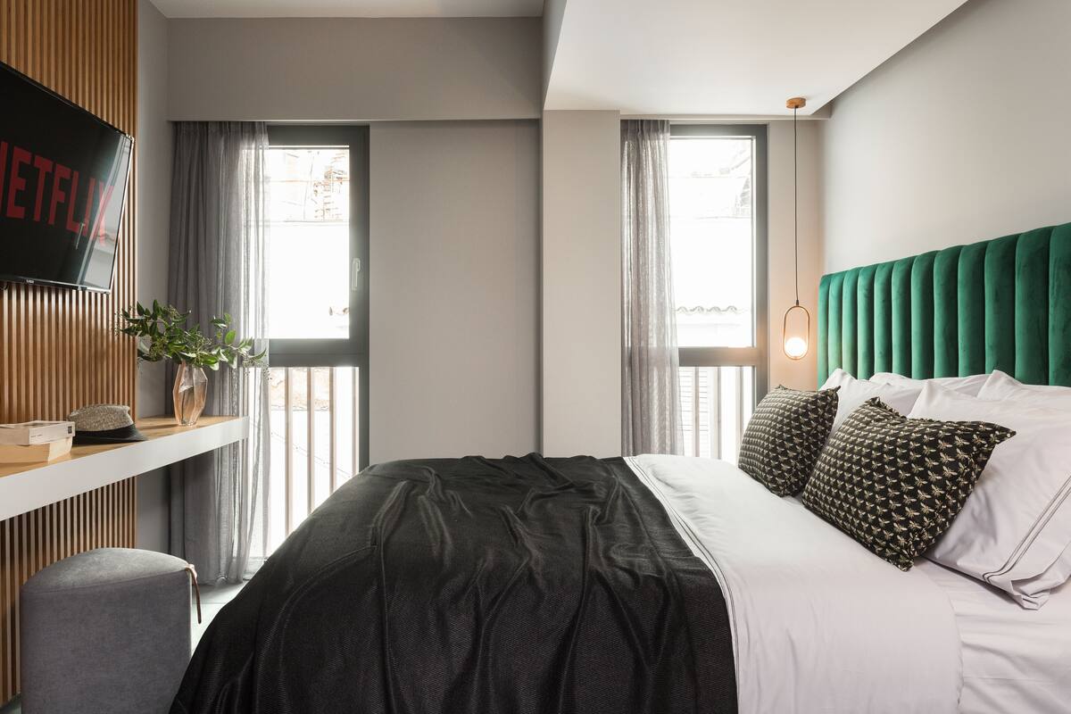 Une chambre moderne avec un grand lit, une tête de lit verte en tissu, un téléviseur fixé au mur qui affiche « NETFLIX », et une fenêtre avec des rideaux.
