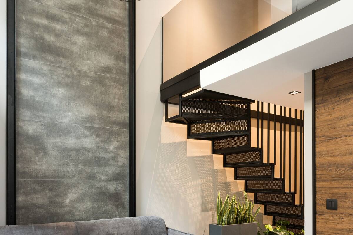 Un salon moderne avec un canapé gris, deux petites tables rondes, un escalier avec des éléments en bois et un luminaire suspendu.