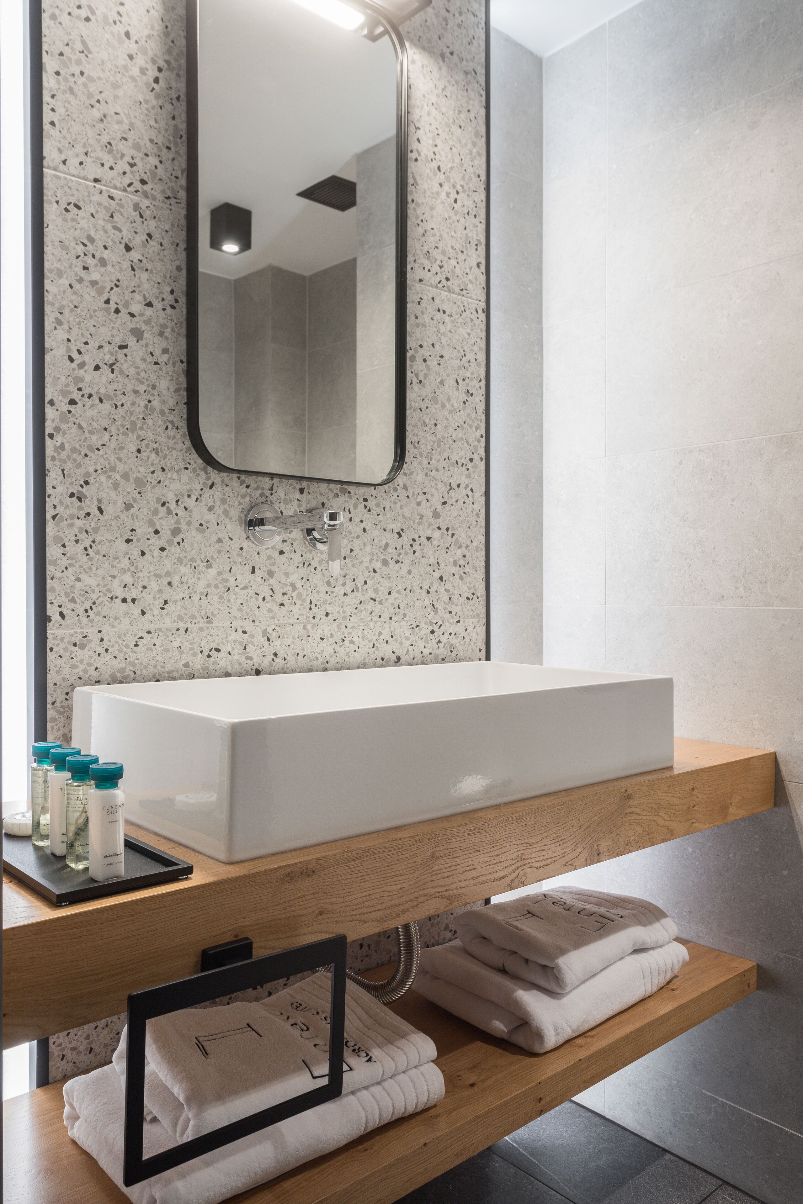 A modern bathroom with a rectangular sink, a mirror, and a wooden shelf with towels and toiletries.