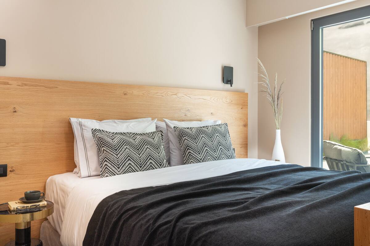 Une chambre à coucher moderne avec une tête de lit en bois, un lit agrémenté d’oreillers à motifs, une table de chevet avec une tasse et un livre, et une vue dégagée sur l’extérieur grâce à une grande fenêtre.